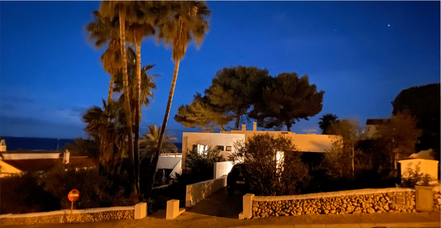 La Villa Caroline au coucher du soleil, une villa blanche à toit plat derrière un mur minorquin en pierre sèche, avec de hauts palmiers et des pins et la mer juste derrière.