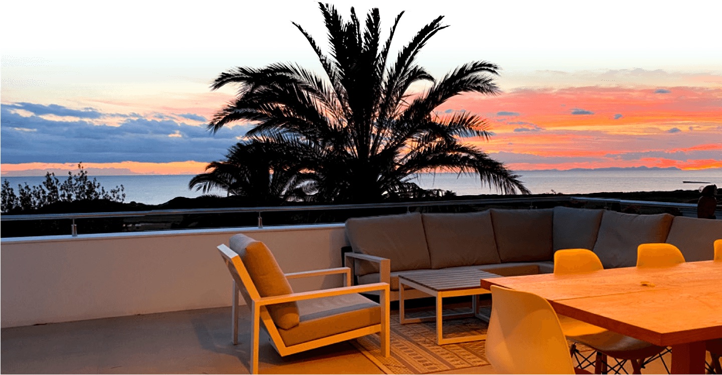 La terrasse de la villa au coucher du soleil, avec un canapé d'angle, une table à manger et un grand palmier se découpant sur un ciel rose et orange au-dessus de la mer.